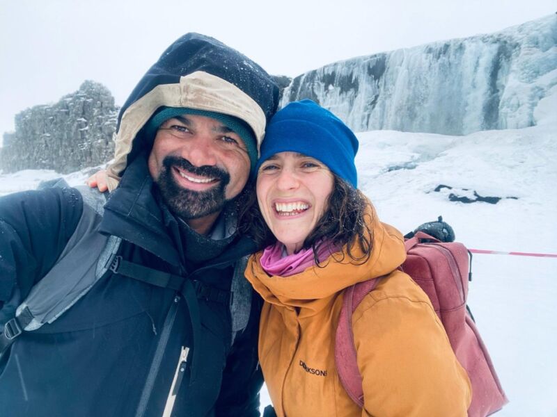 A portrait of a man and a woman, hugging and smiling in the camera. In the background you can see a snow field and a waterfall.